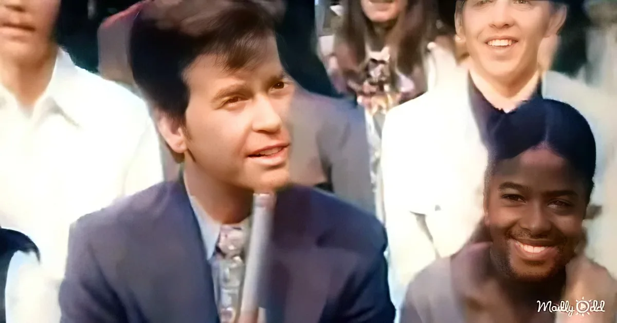 A man in a suit holding a microphone, surrounded by smiling people.