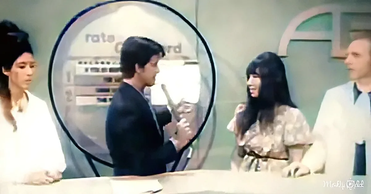 A man in a suit holds a microphone, speaking to a woman with long hair, while others observe in a studio setting.