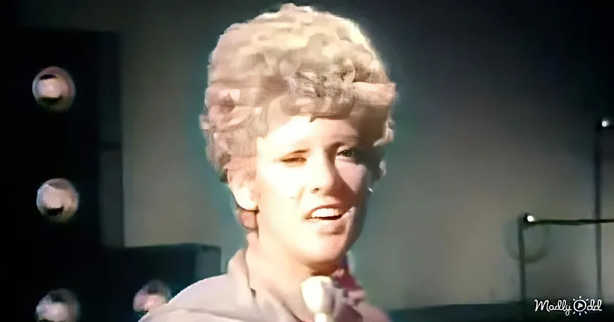A person with curly blonde hair is holding a microphone, possibly singing or speaking.
