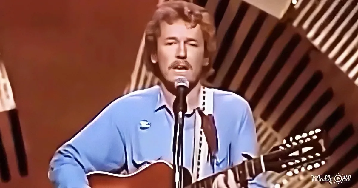 A man in a blue shirt sings into a microphone while playing an acoustic guitar.