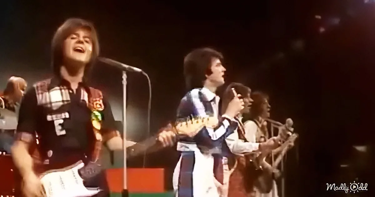 A man in a plaid shirt sings into a microphone on stage, surrounded by other musicians.
