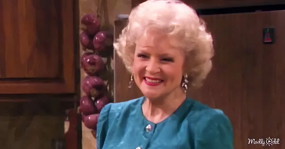 A smiling woman with blonde hair in a teal dress stands in a kitchen with wooden cabinets and a bunch of purple onions.