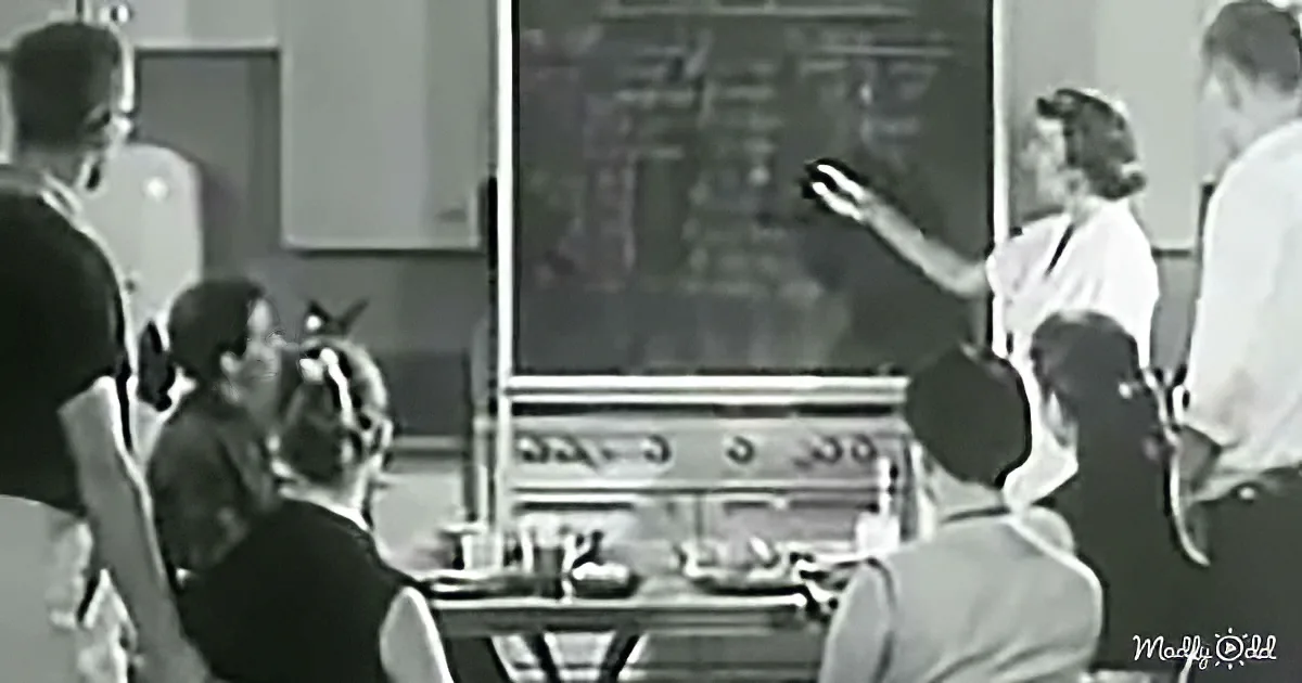 A teacher points at a chalkboard while students sit in a classroom.