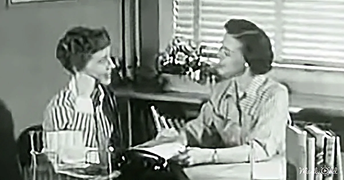 Two women sit at a table, one gesturing while the other listens, in a vintage setting.