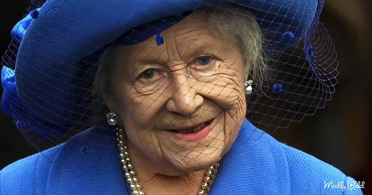 An elderly woman in a blue outfit and hat with a net veil, smiling.