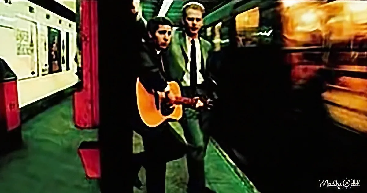 A man in a suit plays guitar on a subway platform, with another man in a green suit standing nearby.