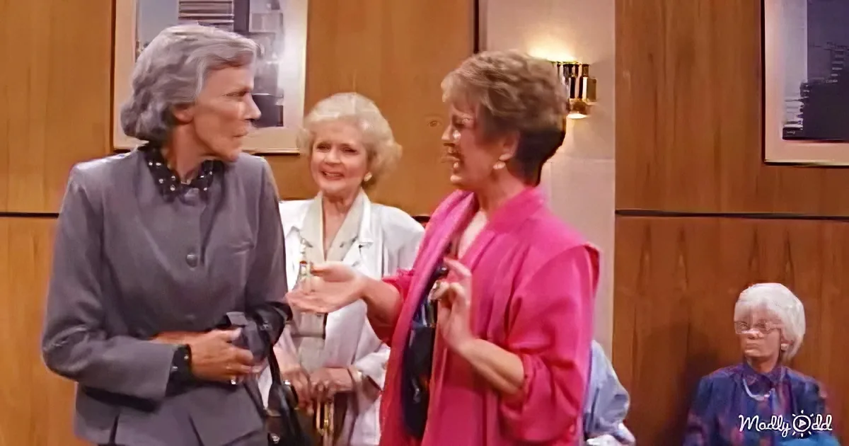 A woman in a pink scarf gestures while talking to another woman in a gray suit in a room with wooden walls.