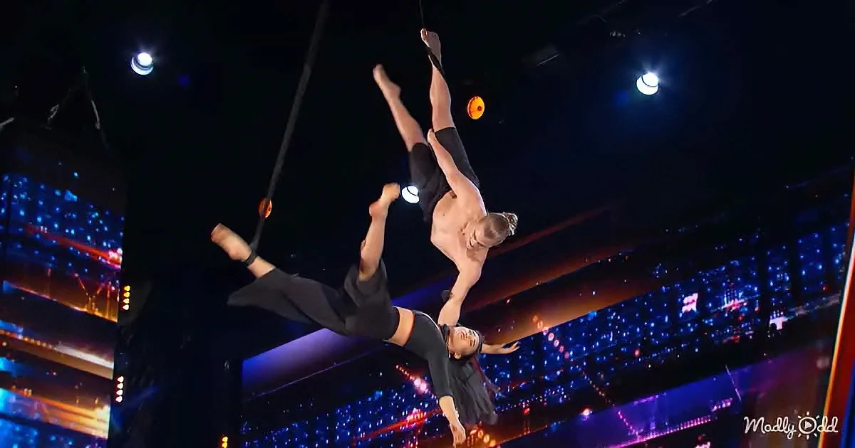 Two acrobats perform a synchronized aerial trick, suspended in mid-air against a dark, illuminated background.