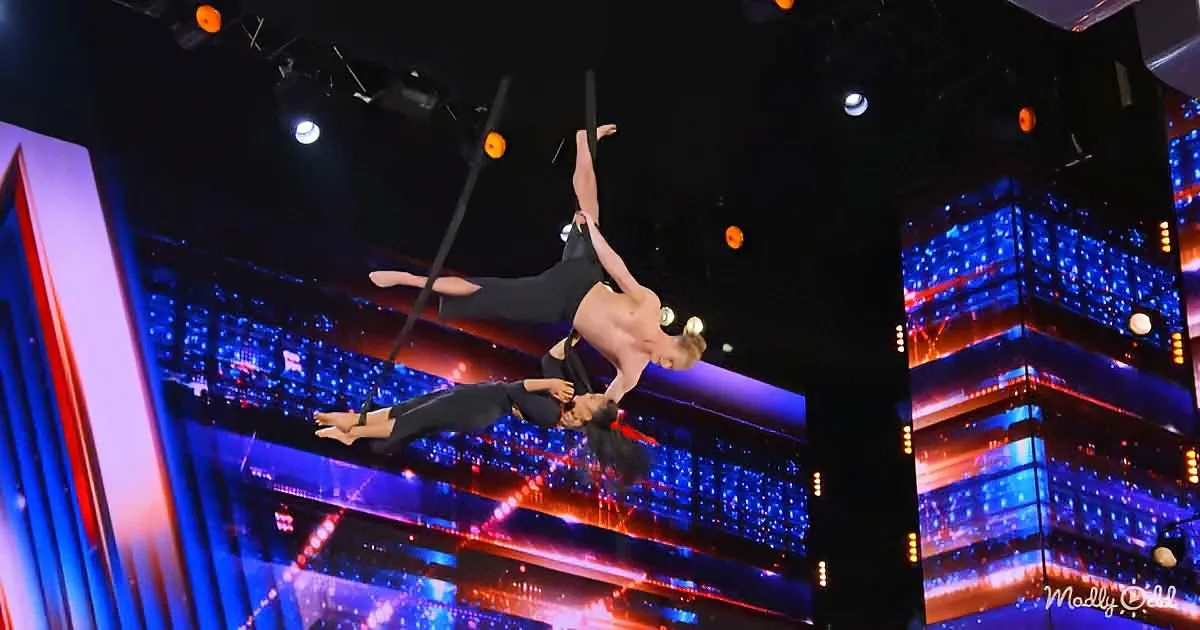 Two acrobats perform a synchronized aerial trick on a stage with vibrant blue and red lighting.