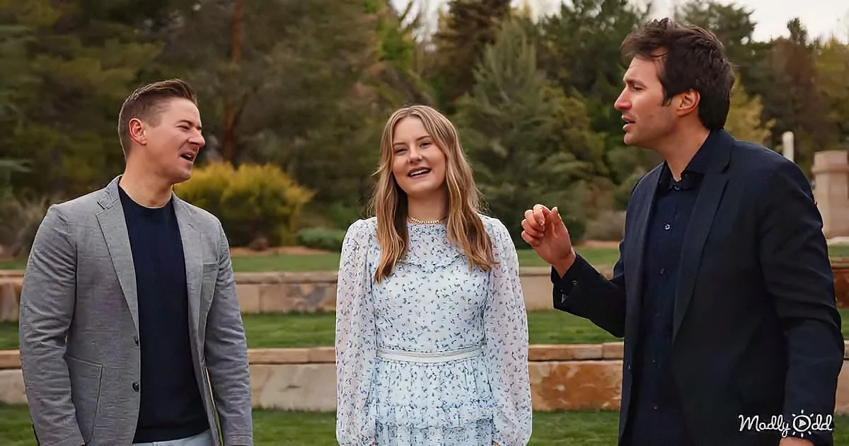A woman in a floral dress smiles as two men, one in a gray suit and the other in a black suit, engage in conversation...