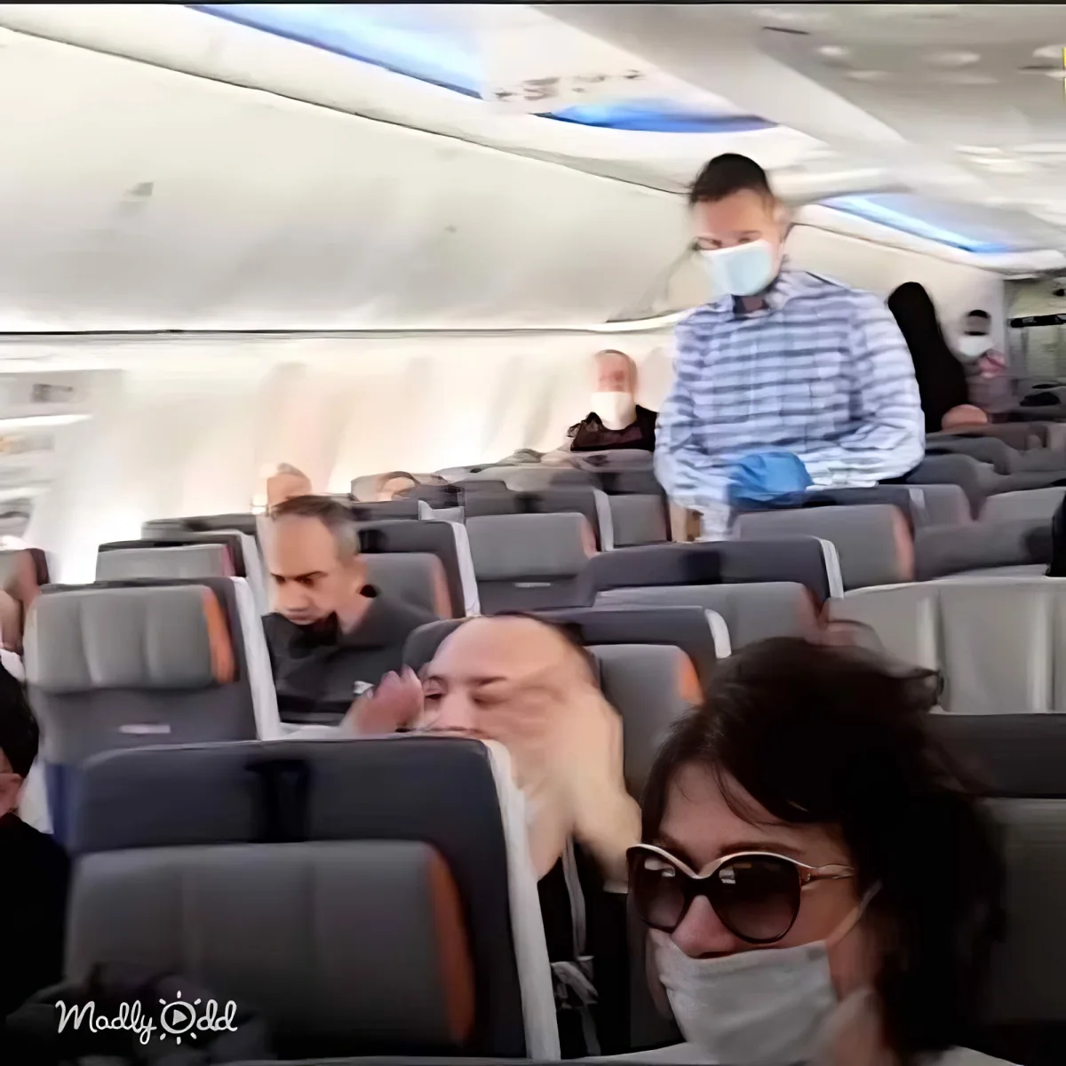 A flight attendant in a blue checkered shirt and gloves walks through an airplane cabin.