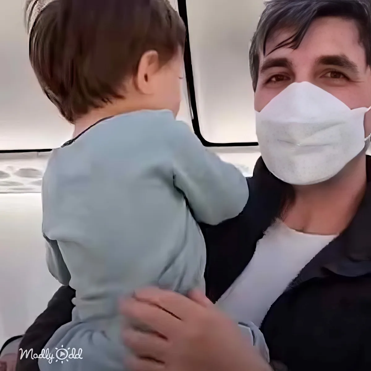 A man wearing a mask holds a child in a light blue outfit, both seated inside a vehicle.