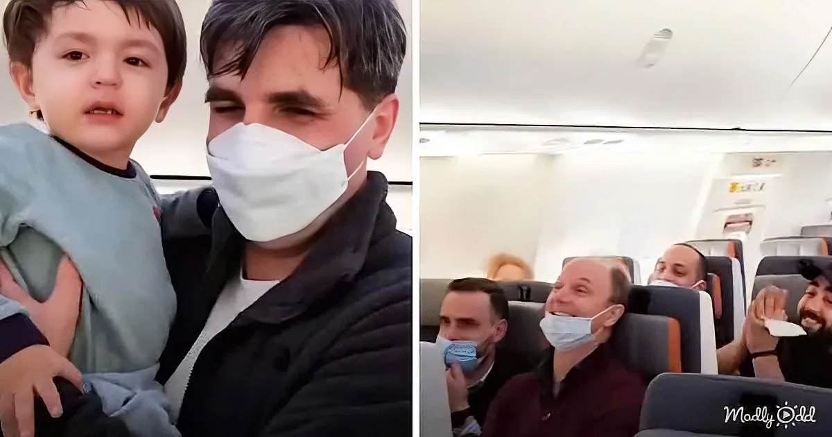 A man wearing a mask holds a child on his lap, while passengers in the background also wear masks on an airplane.