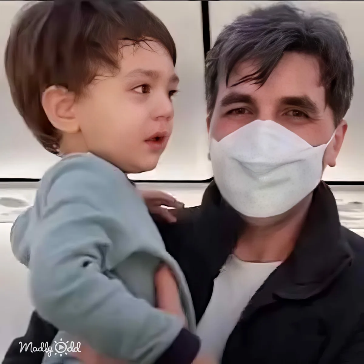 A man wearing a mask holds a child in a light blue shirt, both looking to the side.