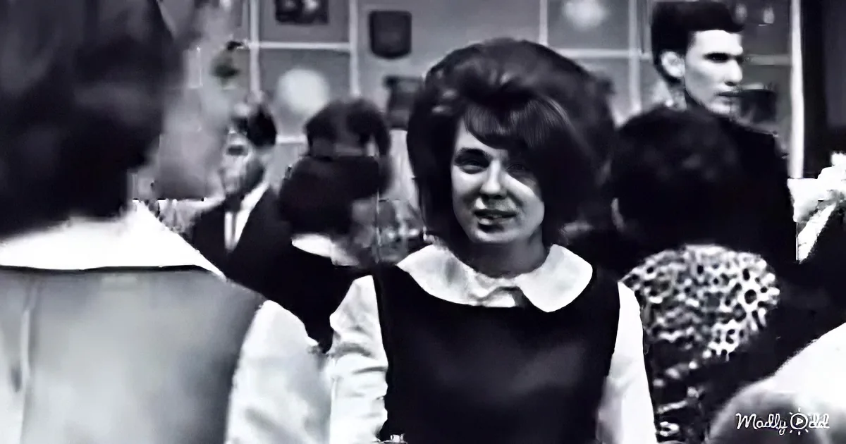 A young girl in a black dress with a white collar is smiling and raising her hand in a classroom setting.