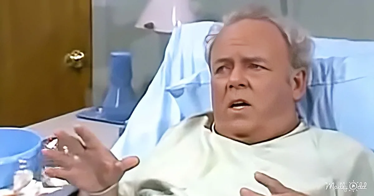 A man in a hospital bed, wearing a white gown, gestures with his hands while speaking.