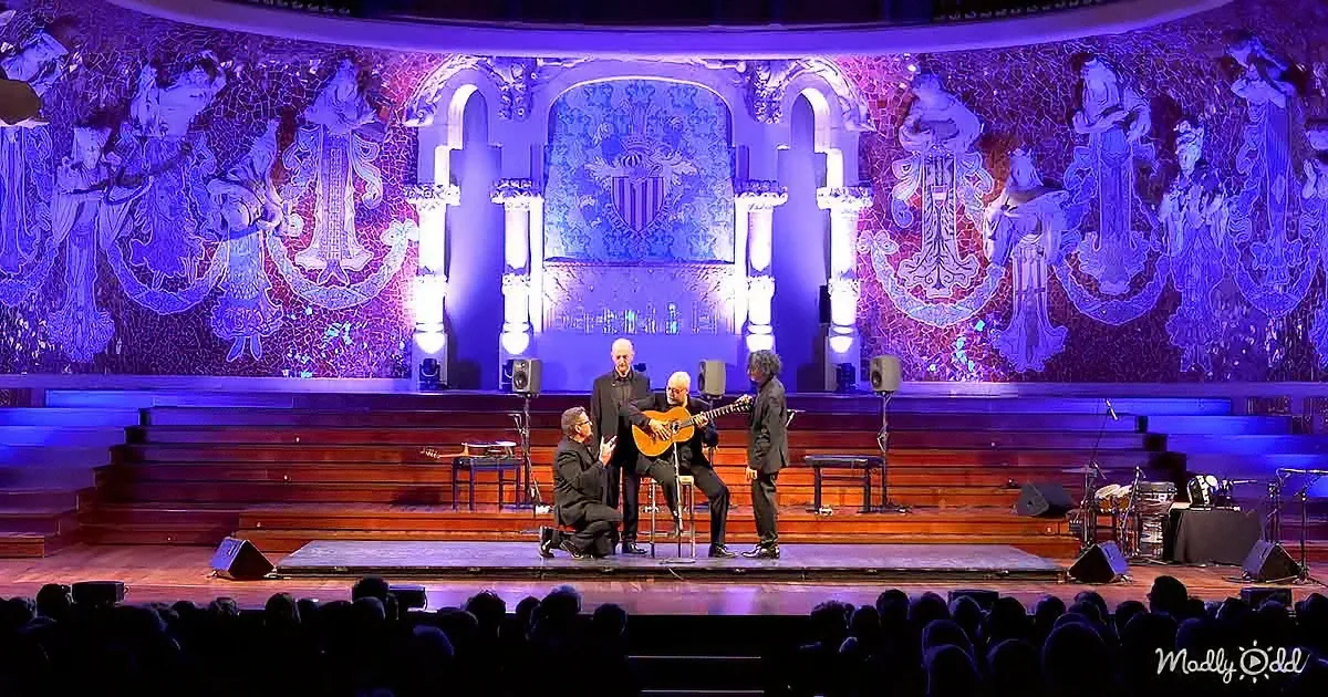 A guitarist performs on stage with two other musicians, surrounded by ornate blue lighting and intricate wall designs.