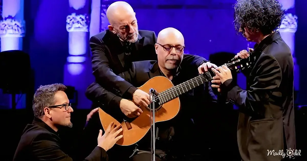 A man in a black suit plays an acoustic guitar with another man assisting, while a third man in a suit looks on.