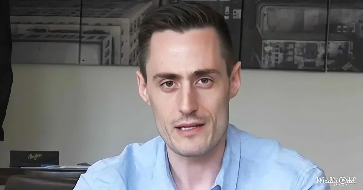A man with short brown hair, wearing a light blue shirt, is seated in a room with a white wall and a blackboard in the...