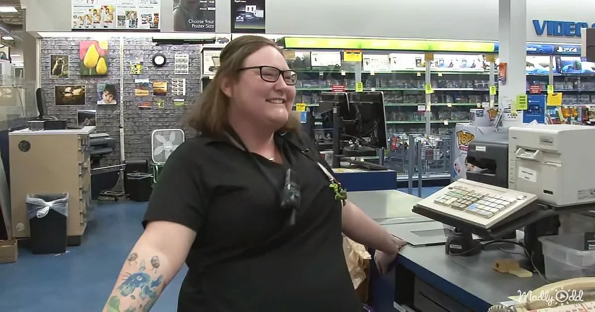 A smiling woman with tattoos stands behind a counter in a store, surrounded by shelves and equipment.