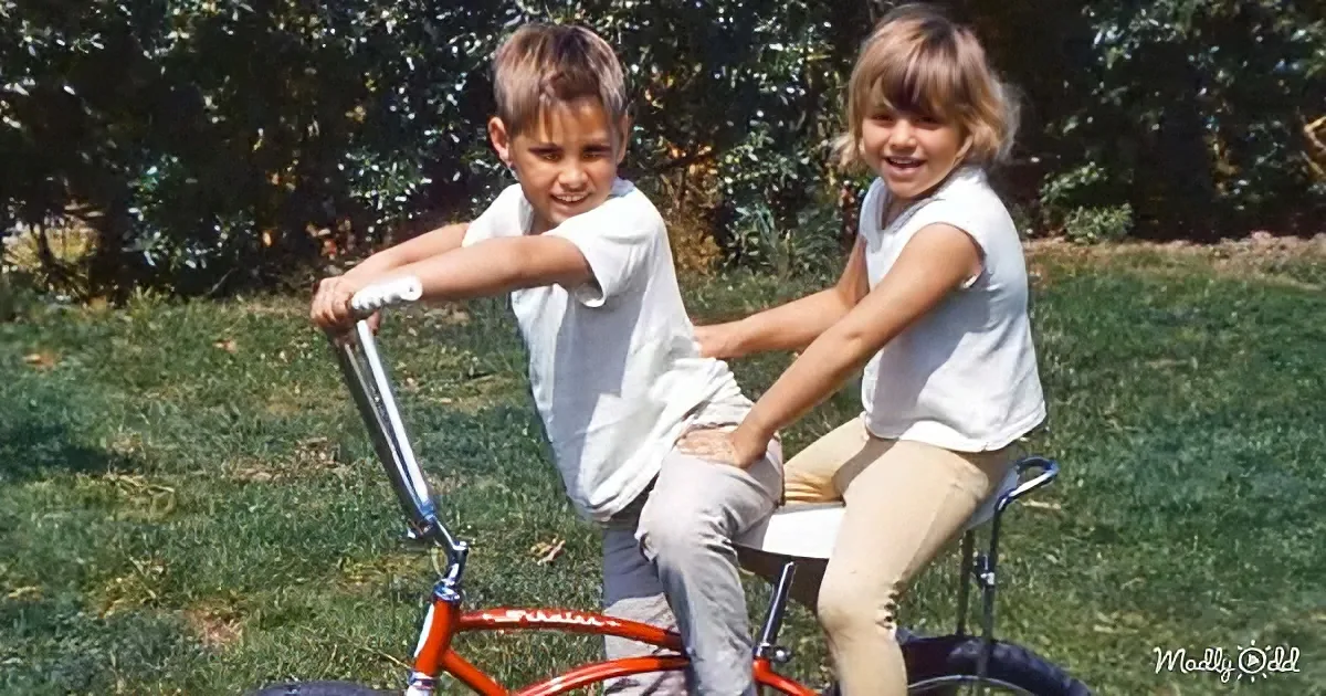 Two children, a boy and a girl, sit on a red bicycle in a grassy area, smiling.