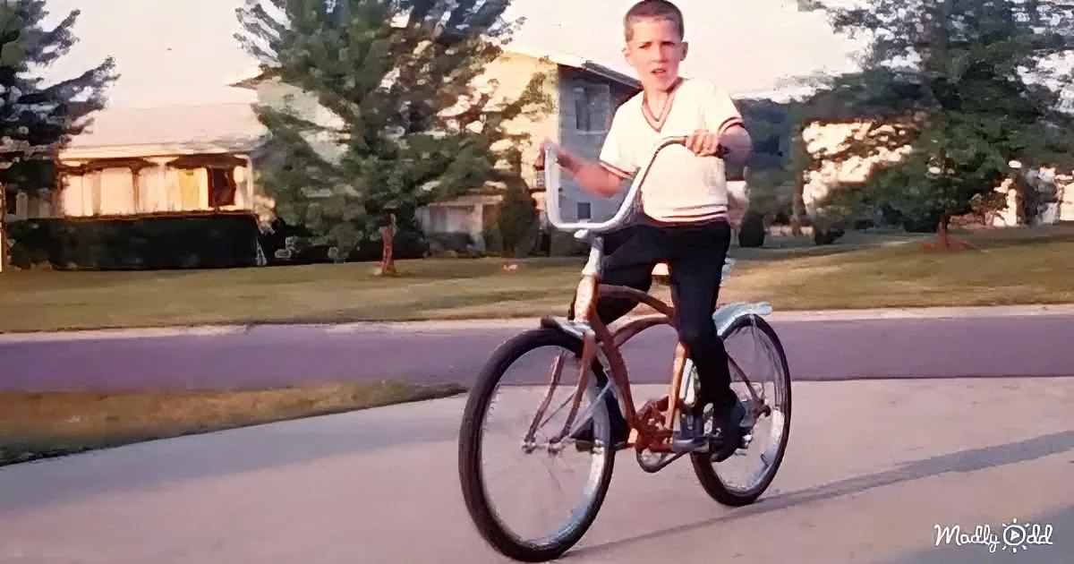 A young boy rides a bicycle on a suburban street, wearing a white shirt and dark pants.