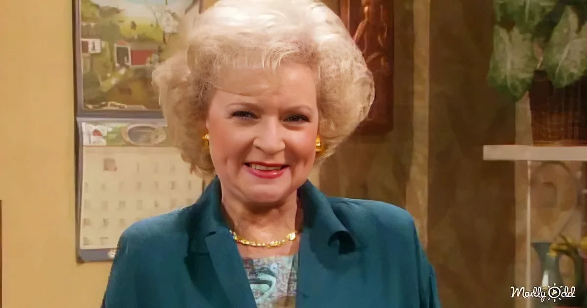 A woman with blonde, curly hair, wearing a teal blazer and a gold necklace, smiles warmly in a room with a calendar and...