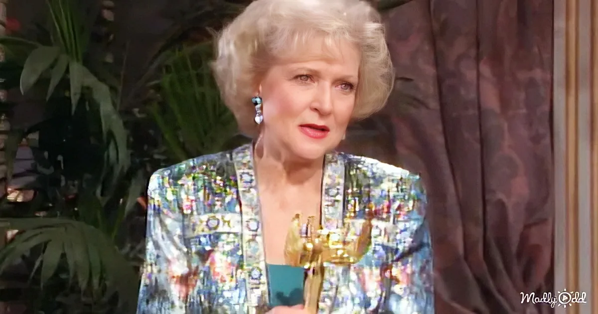 A woman with blonde hair, wearing a colorful patterned jacket, holds a gold award.
