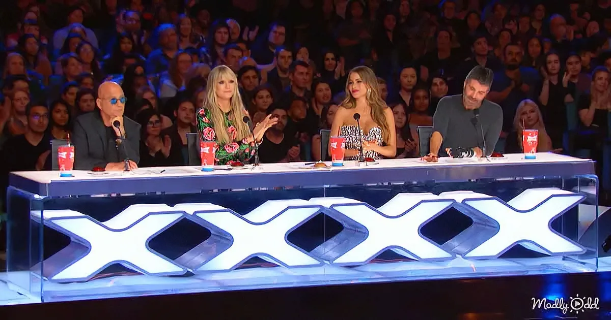 Judges on a panel with "XXXX" sign, audience in the background.