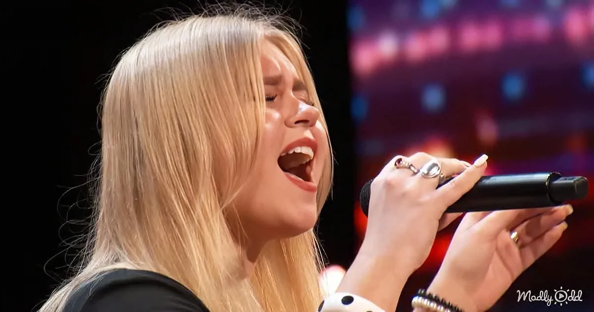 A woman with long blonde hair sings passionately into a microphone on stage.