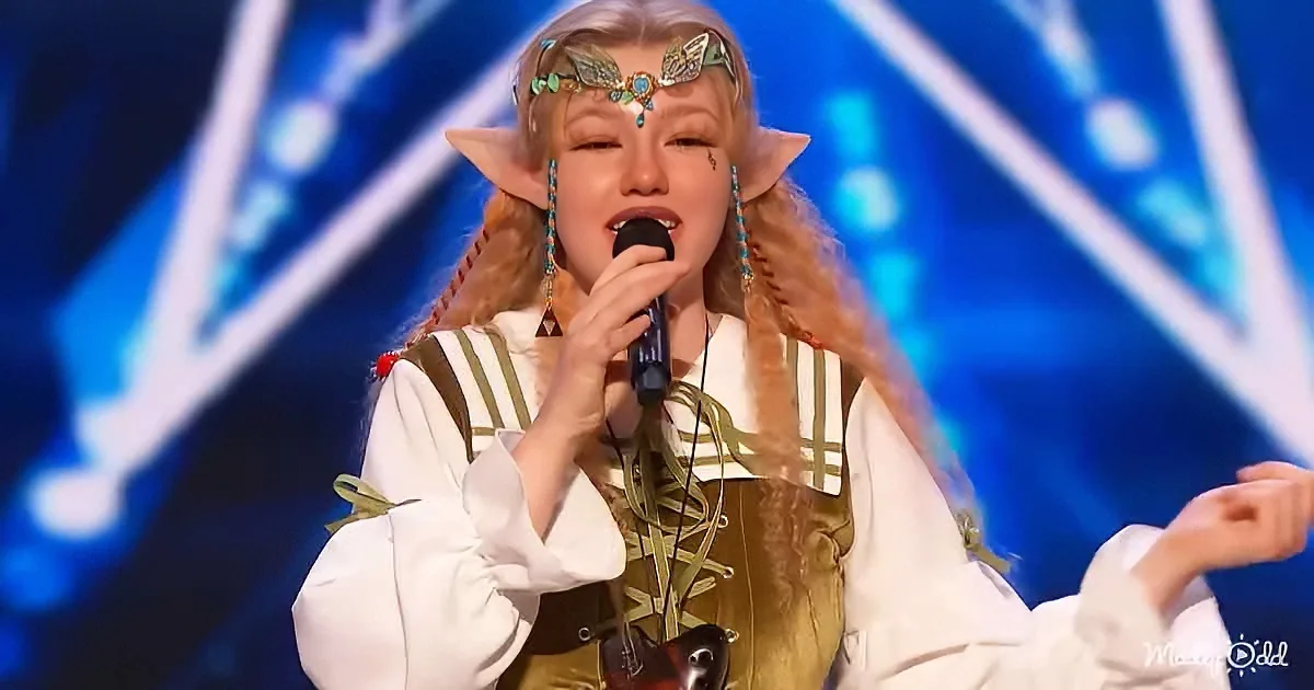 A person in an elf costume sings into a microphone on a stage with blue lights.