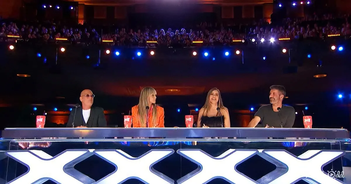 Four judges sit at a panel with a large "X" logo, facing a dimly lit audience in a theater.
