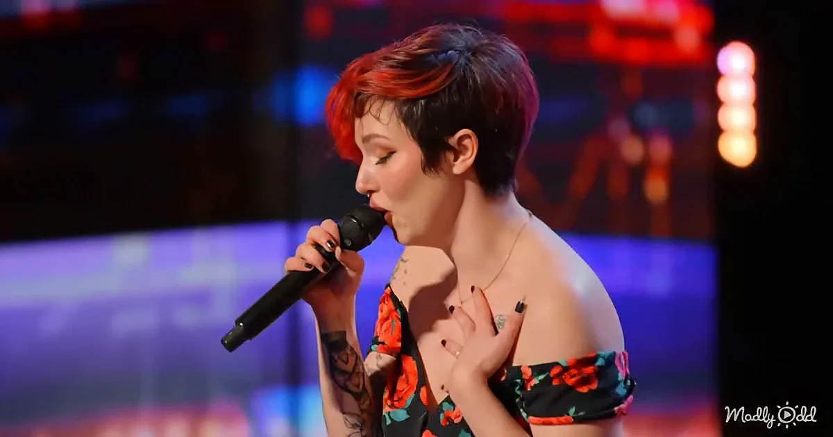 A person with red and black hair sings into a microphone, wearing a floral dress and a necklace.