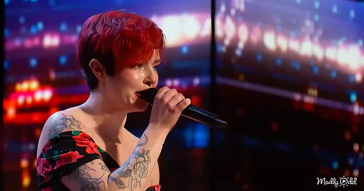 A person with red hair and tattoos sings into a microphone on a stage with colorful lights.