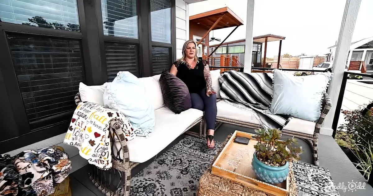 A woman sits on a wicker sofa adorned with pillows and blankets on a cozy patio.