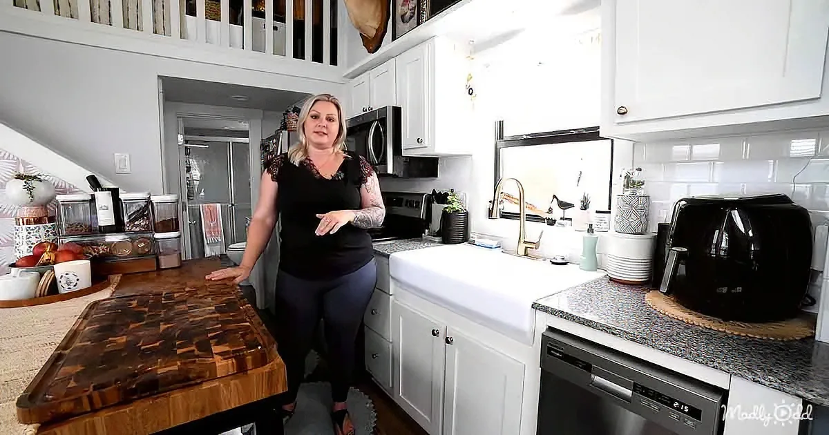 A woman stands in a modern kitchen with white cabinets, granite countertops, and a large farmhouse sink.