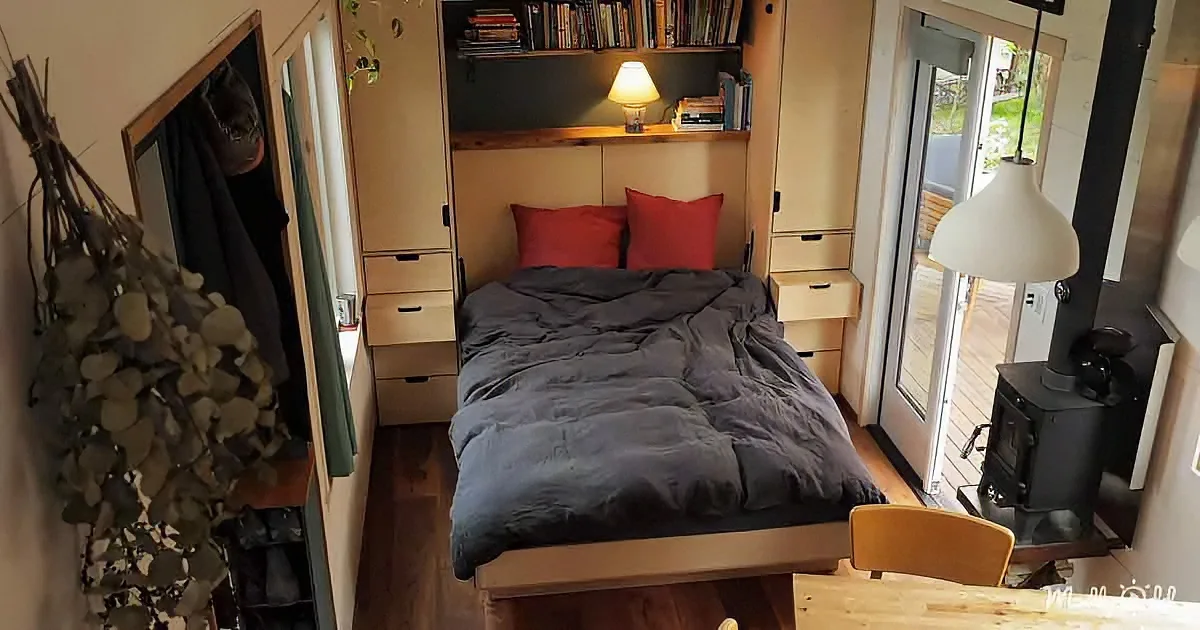 A cozy bedroom with a bed, red pillows, wooden shelves, and a sliding glass door leading outside.