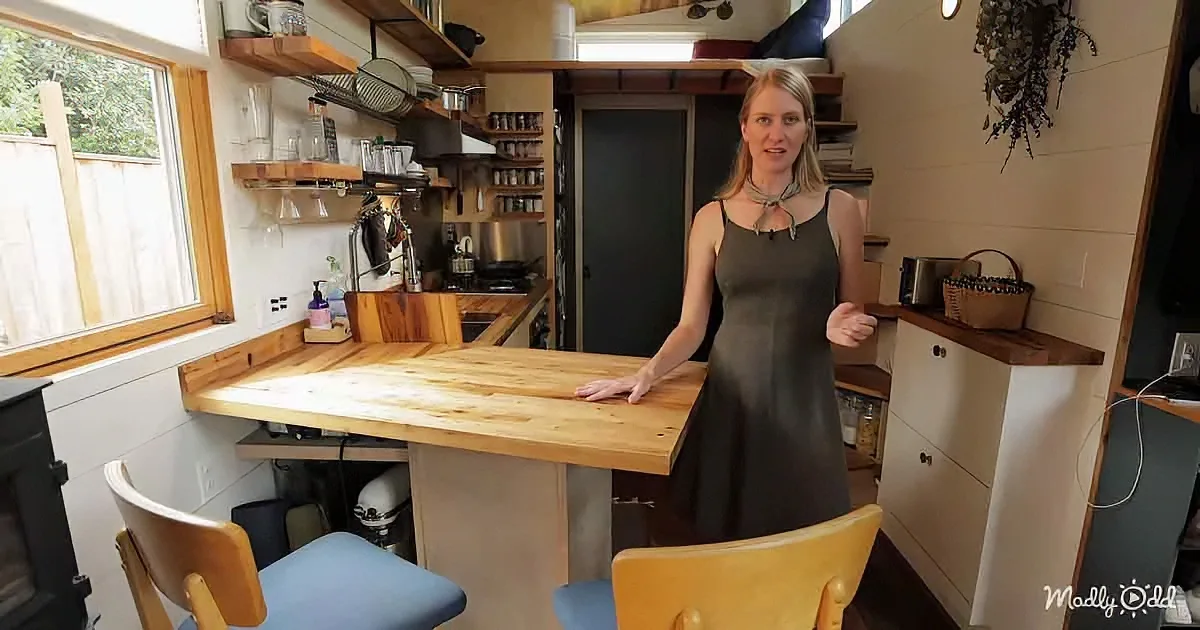 A woman stands in a cozy, compact kitchen with wooden shelves, a large island, and a window overlooking a fence.