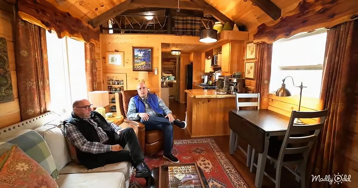 Two men sit in a cozy cabin interior with wooden walls, furniture, and a warm, rustic ambiance.