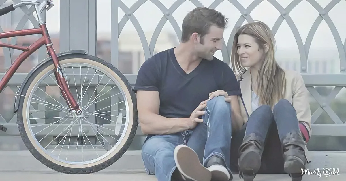 A couple sits on a bench, smiling, with a red bicycle beside them.