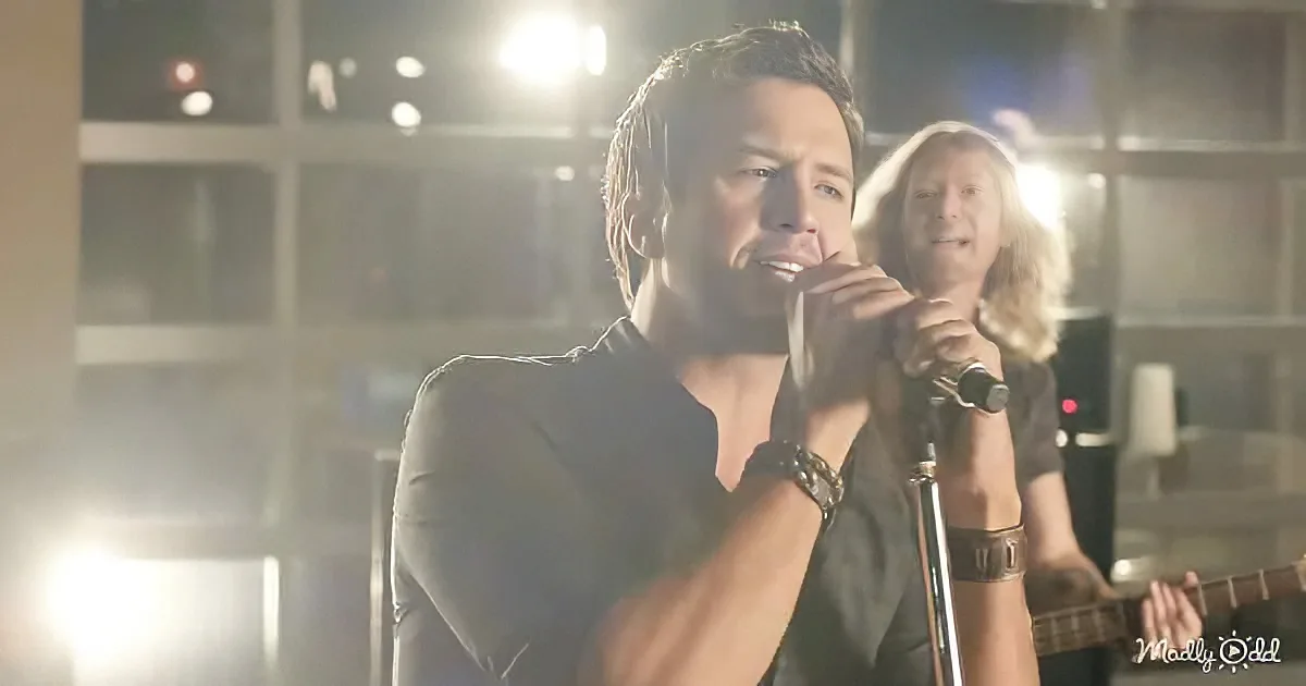 A man in a black shirt sings into a microphone, with another person playing guitar in the background.