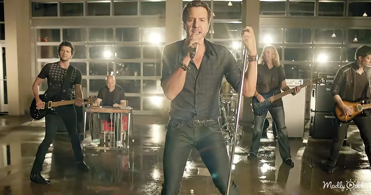 A man in a black shirt sings into a microphone, surrounded by band members playing guitars and drums in a dimly lit room.