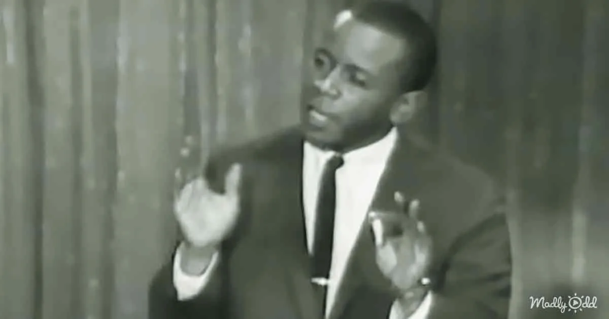 A man in a suit and tie gestures with his hands, standing in front of a curtain.