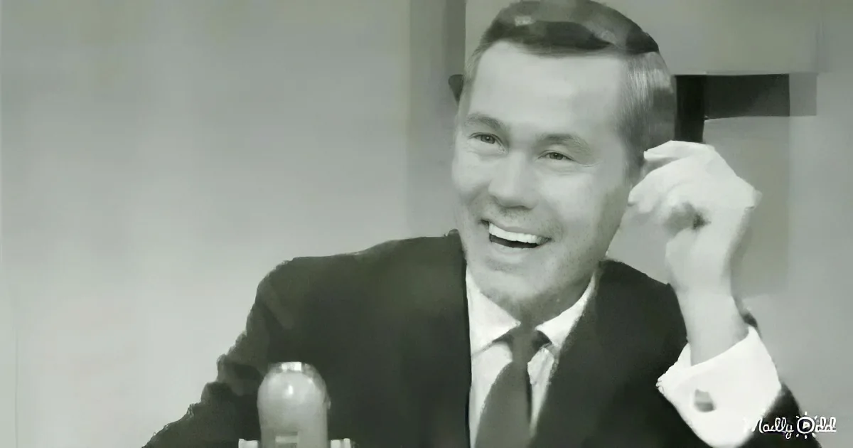 A man in a suit and tie is smiling, holding a microphone, and appears to be speaking or presenting.