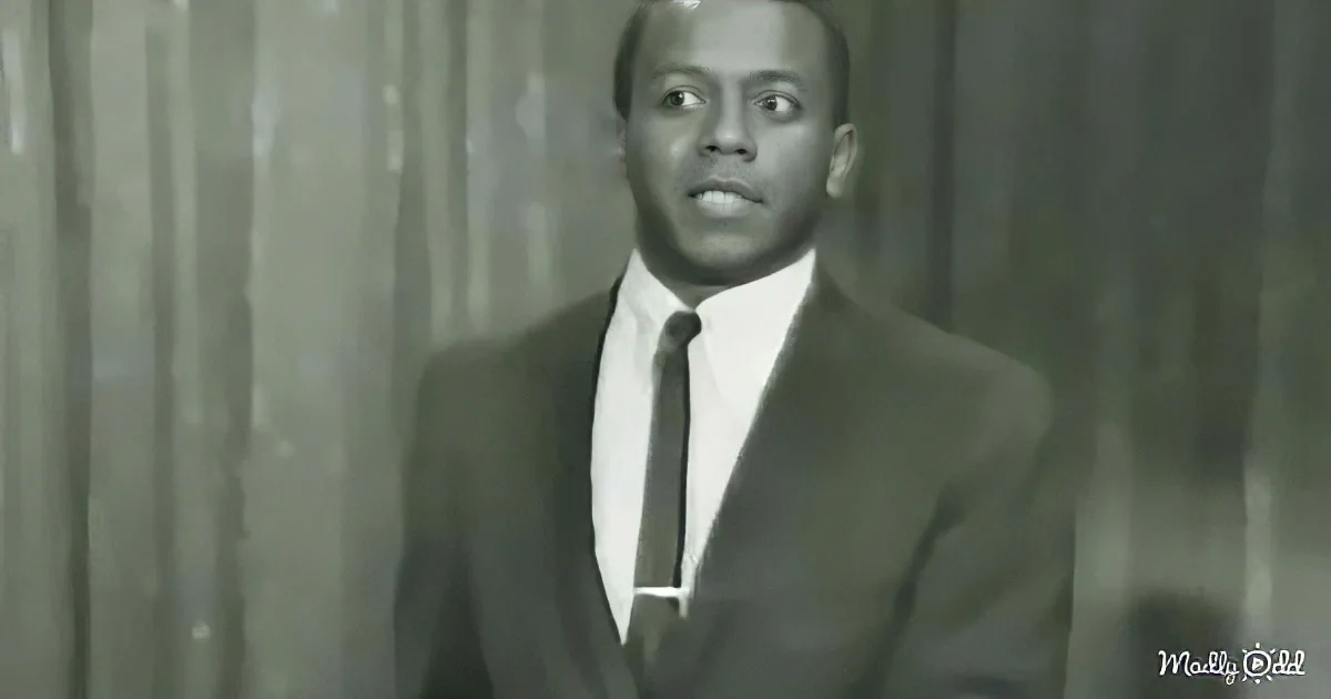 A man in a suit and tie stands against a curtain backdrop, looking slightly to the side.