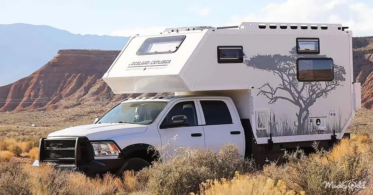 A white Overland Explorer truck camper is parked in a desert landscape with a tree silhouette on its side.