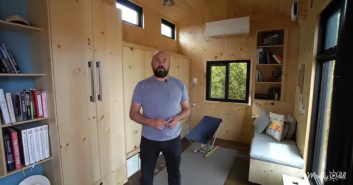 A man stands in a cozy, compact room with wooden walls, a small bed, bookshelves, and a chair.