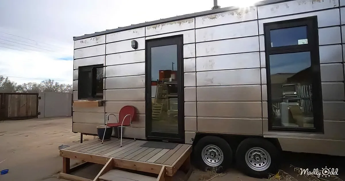 A silver tiny house on wheels with a wooden deck and red chair, set against a cloudy sky.