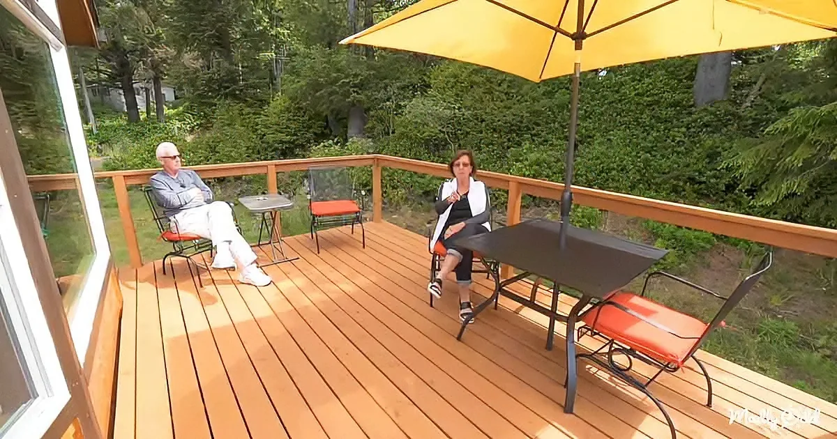 A man and woman sit on a wooden deck surrounded by greenery, under a yellow umbrella.
