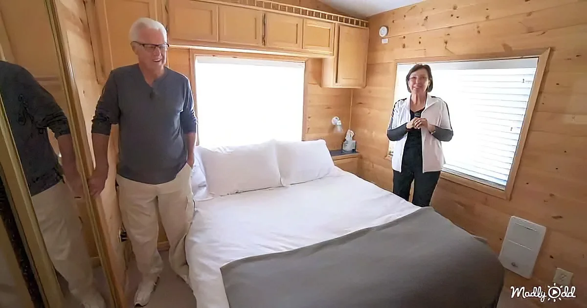 A man and woman stand in a cozy, wooden cabin bedroom with a neatly made bed and windows.